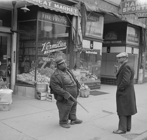 Two men stand on a sidewalk in front of a meat market and produce store. One man, dressed in work clothes and holding a broom, faces another man wearing a long coat and cap. Produce baskets are visible outside the store.