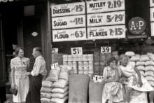 Two women and a man stand and talk outside a grocery store, while two girls sit on sacks of flour. Storefront windows display prices for salad dressing, margarine, sugar, milk, flour, and corn flakes.