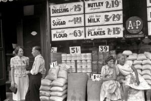 Two women and a man stand and talk outside a grocery store, while two girls sit on sacks of flour. Storefront windows display prices for salad dressing, margarine, sugar, milk, flour, and corn flakes.