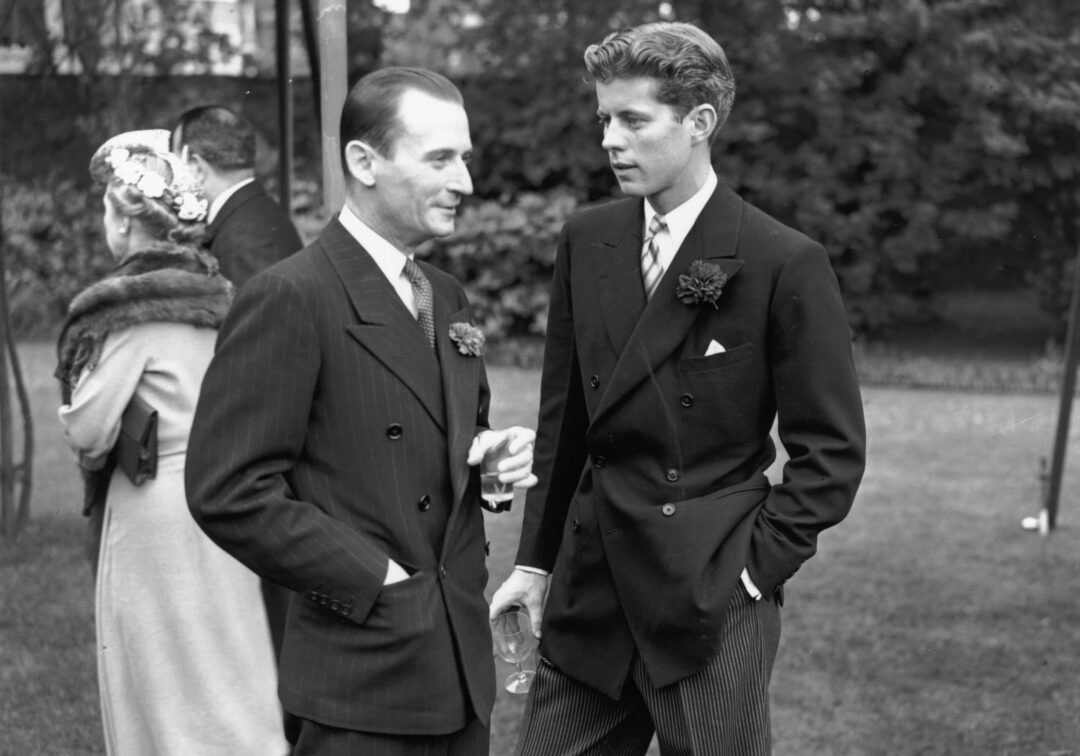 Two men in suits have a conversation outdoors; one holds a drink. In the background, a woman in a fur-collared coat and hat stands with another man, with trees and a lawn visible. The image is black and white.