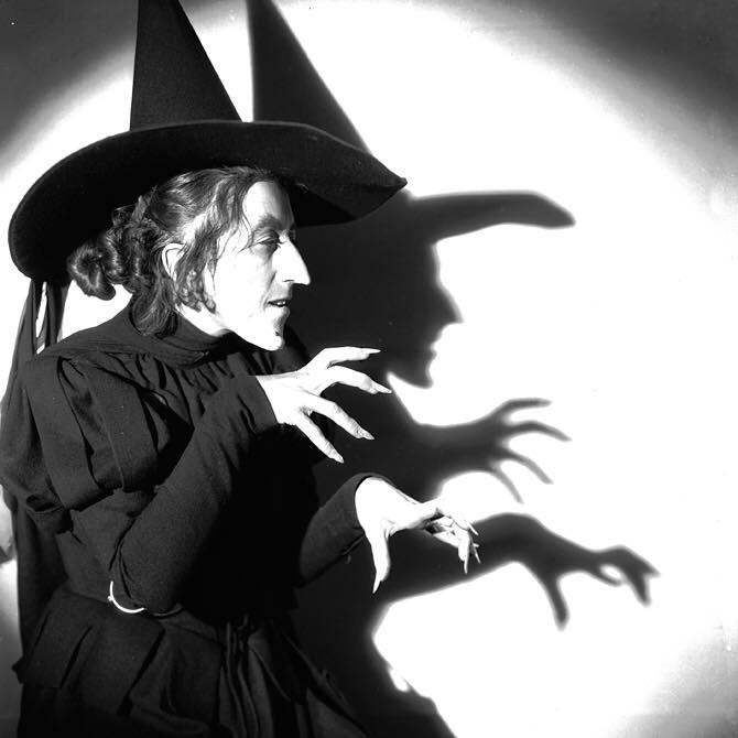 A person dressed as a classic witch with a pointed hat and long, claw-like fingers poses dramatically, casting a creepy shadow with outstretched hands on a white wall behind them.