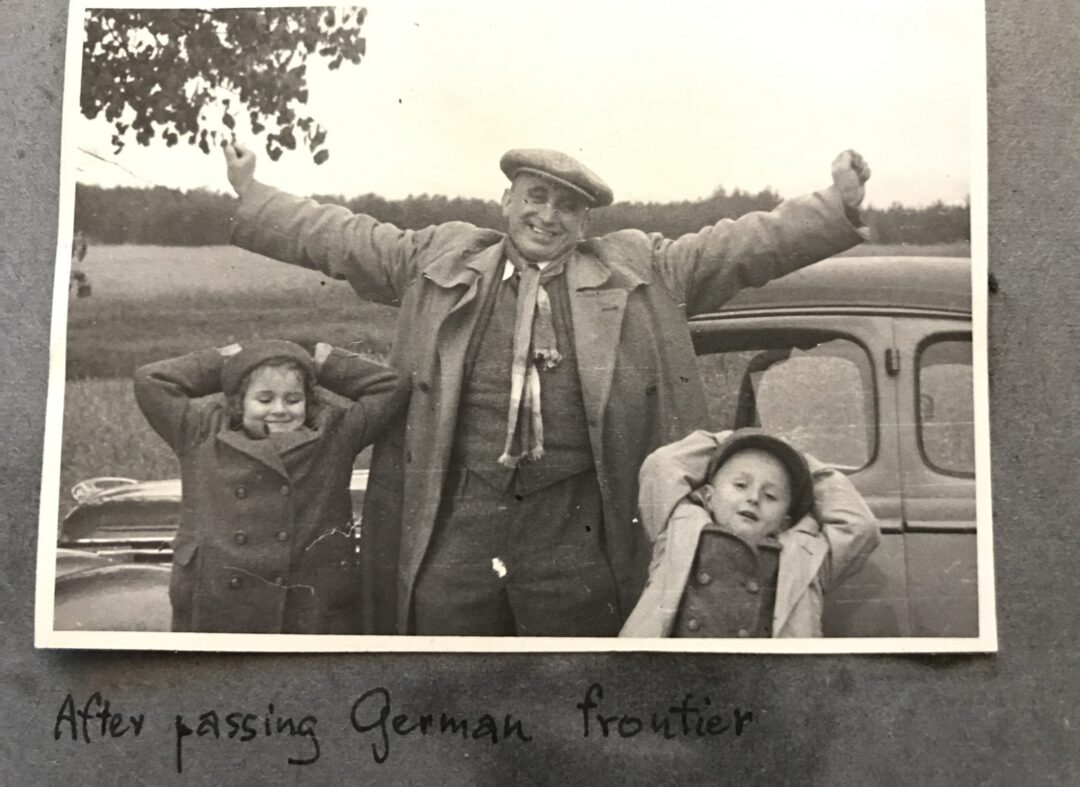 A black and white photo shows a smiling man with arms outstretched standing between two children leaning back, hands behind their heads, in front of a car. Handwritten text below reads: "After passing German frontier.