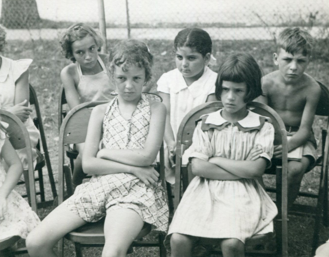 Five children sit outdoors on chairs, all looking serious or unhappy, with arms crossed or hands in laps. A fence and grass are visible in the background. The photo is in black and white.