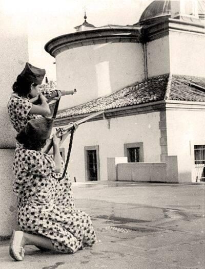 Two women in patterned dresses, one kneeling and one standing, aim rifles from behind a stone structure. The background features a round building with a tiled roof and dome. The scene appears historical and tense.