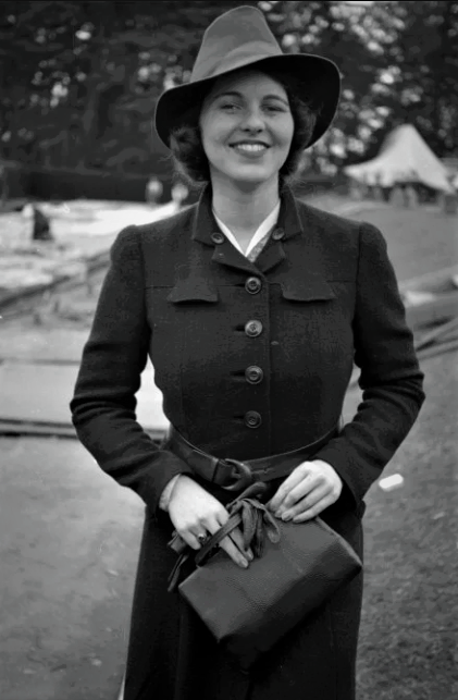 A woman in a dark coat and wide-brimmed hat smiles while holding a handbag outdoors, with trees and tents visible in the background. The photo appears to be from a past era, possibly the mid-20th century.