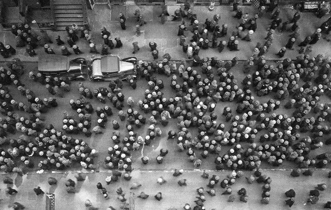 Aerial view of a crowded city street, filled with people wearing hats and walking in various directions. Two cars are parked along the street, surrounded by the bustling crowd.