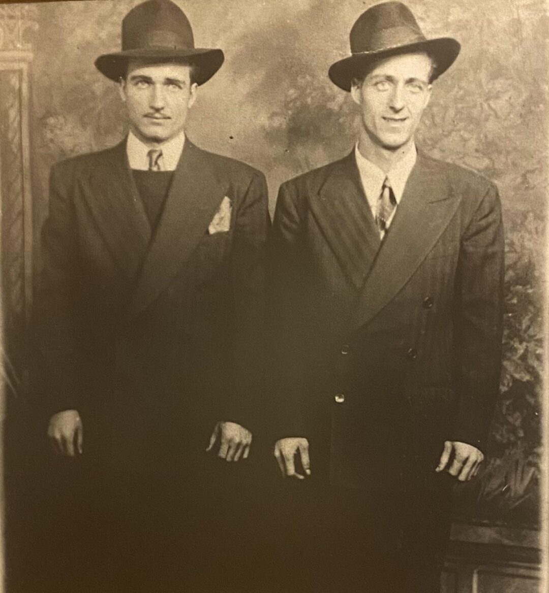 Two men in dark suits and wide-brimmed hats stand side by side, facing the camera. Both wear ties and have serious expressions. The photo is sepia-toned and appears old-fashioned, with a painted studio backdrop.