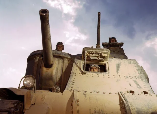 Low-angle view of a dusty World War II tank with two soldiers visible through hatches, against a partly cloudy sky. The tank’s large gun barrels and armored exterior dominate the foreground.