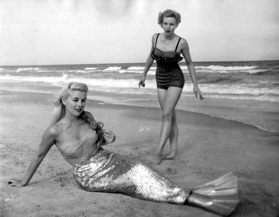 Two women pose on a beach; one is sitting on the sand wearing a shiny mermaid tail costume, while the other stands nearby in a striped vintage swimsuit. Ocean waves and sky are visible in the background.