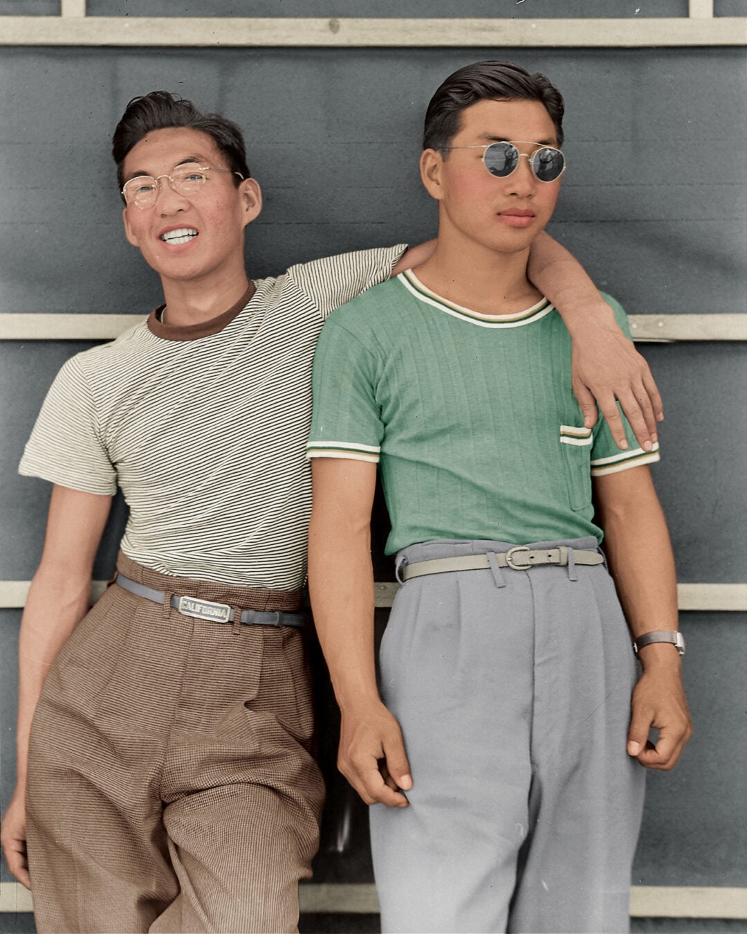 Two young men stand side by side, one with his arm around the other. Both wear high-waisted trousers and short-sleeved shirts. One smiles, wearing glasses, while the other looks serious in sunglasses, against a slatted wall background.