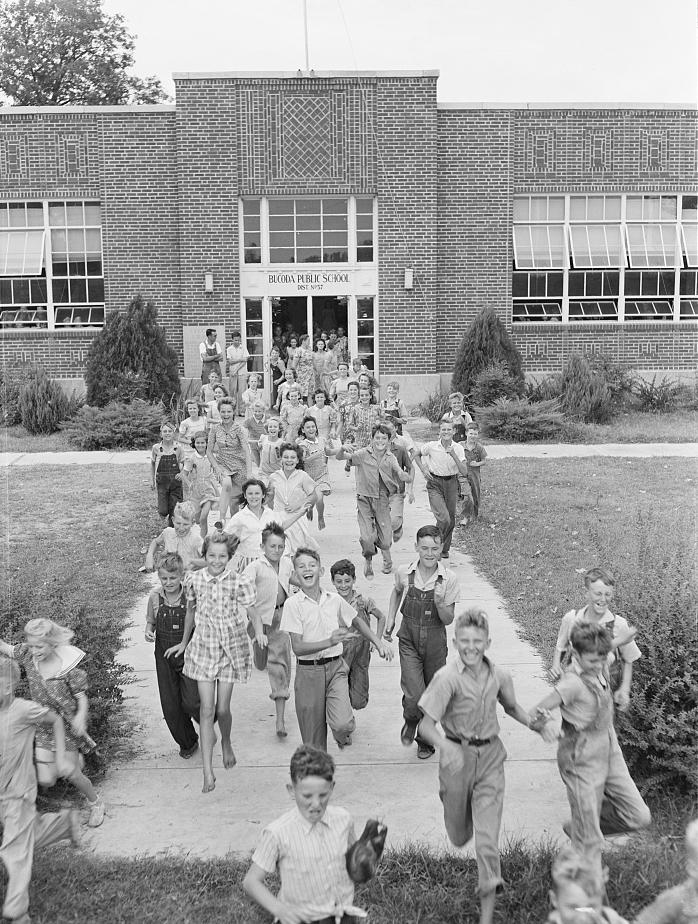 A large group of smiling children run out of the entrance of a brick school building labeled "Bloom Public School" on a sunny day, excitedly heading down the front walkway.
