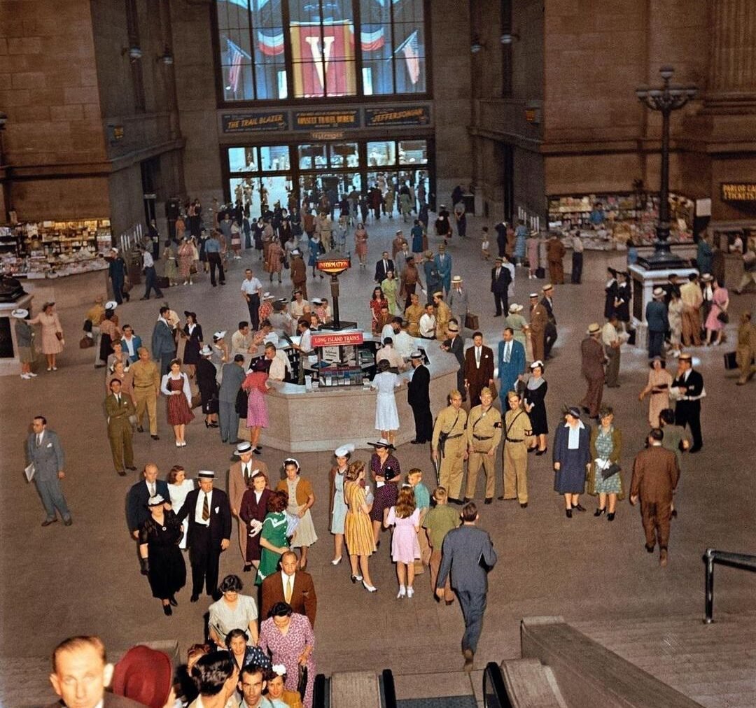A busy train station in the 1940s with people in vintage clothing, some in military uniforms, walking or standing in groups. An American flag hangs above the entrance, and shops line the sides.