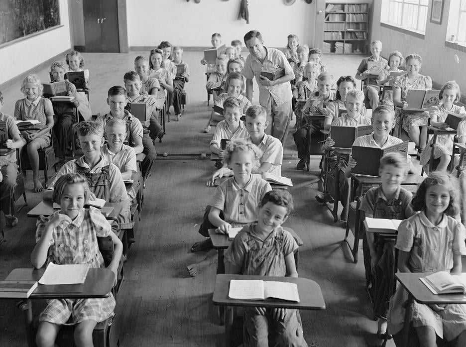 A black-and-white photo of a classroom filled with smiling children seated at desks, facing the camera. An adult stands in the back of the room. The students have books and papers on their desks.