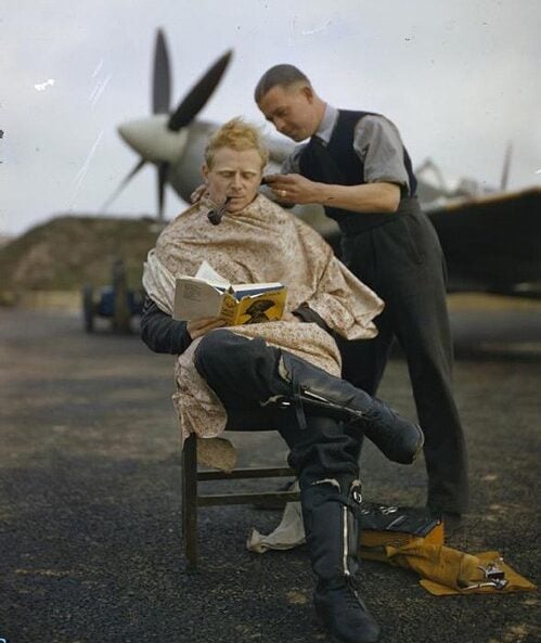 A man sits outdoors getting a haircut while reading a book and smoking a pipe. An airplane propeller and part of a plane are visible in the background, suggesting an airfield setting.