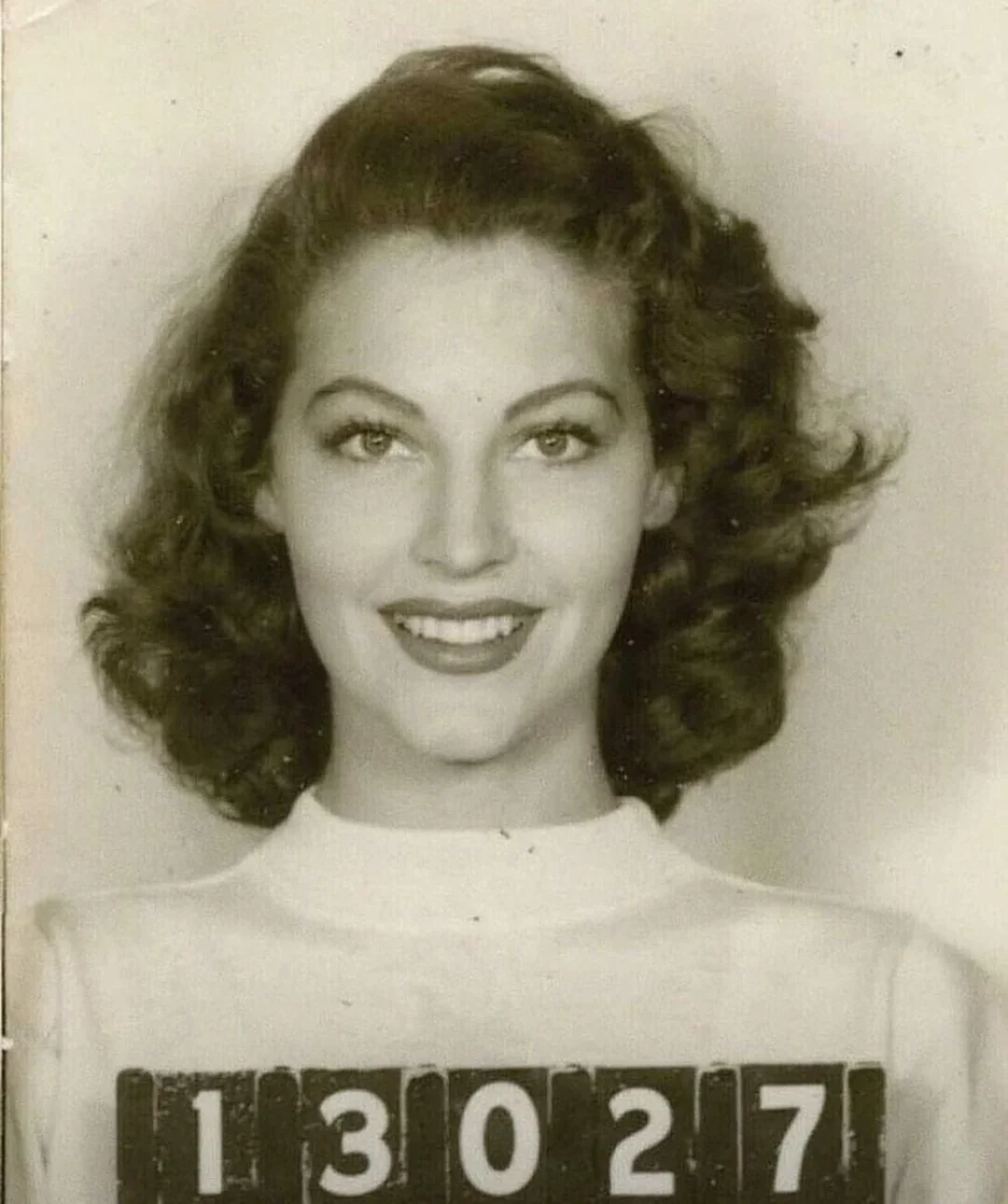 A black and white vintage photo of a woman with wavy hair, smiling, holding a placard with the number 13027, as if for a mugshot.