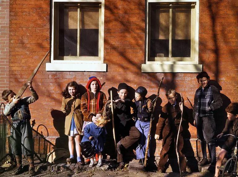 Eight children dressed in winter clothes stand and sit against a brick wall with two windows, holding sticks and smiling, in a sunny outdoor scene. Shadows of tree branches fall across the group.