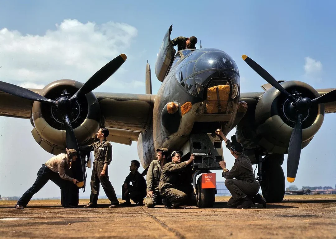A group of men work on or inspect a World War II-era military bomber aircraft parked on a runway under a partly cloudy sky. Some are crouching while others stand near the plane’s nose and propellers.