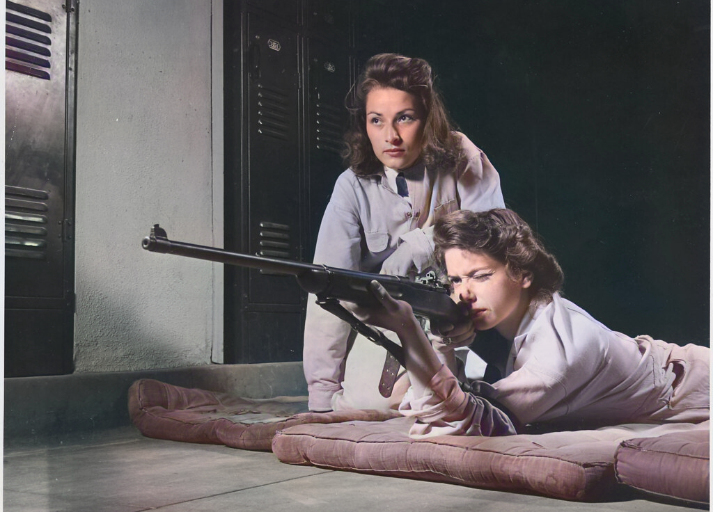 Two women in 1940s clothing practice shooting; one lies prone aiming a rifle while the other kneels beside her, watching attentively. They are indoors on mats near lockers.