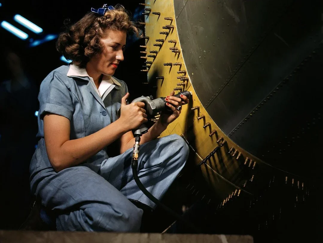 A woman in blue coveralls uses a power tool to work on the fuselage of an aircraft, surrounded by rows of rivets. The scene is well-lit, highlighting her focus and the metallic surface.