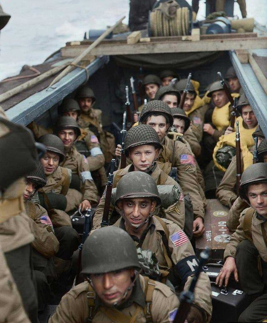 A group of World War II soldiers in uniform and helmets sit closely together in a landing craft, holding rifles, preparing for deployment. Most have serious expressions; the American flag patch is visible on their sleeves.