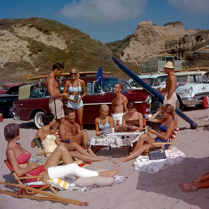 A group of people in swimsuits relax on a sandy beach near vintage cars and a surfboard, some sitting in chairs, others standing and chatting, with rocky cliffs in the background under a partly cloudy sky.