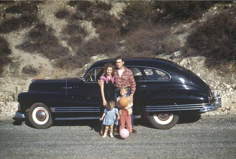A family of four—two adults and two young children—stand in front of a classic dark-colored car on a roadside, with dry, rocky hills in the background. The children are holding colorful balls.