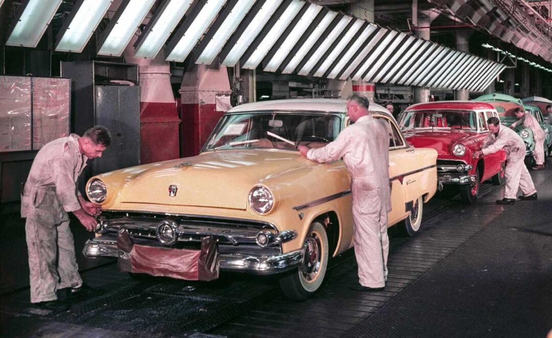 Workers in white coveralls assemble classic cars, including a yellow Ford, on an automotive factory production line under bright overhead lights.