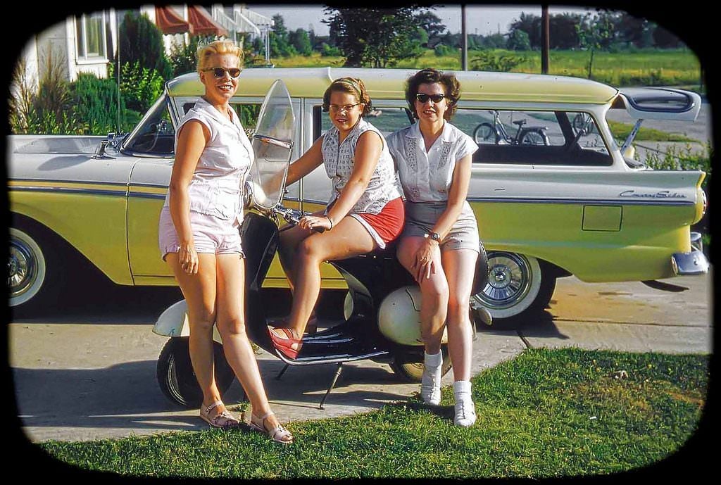 Three women in vintage summer clothing pose with a scooter in front of a yellow station wagon, smiling outdoors on a sunny day with green grass and trees in the background.