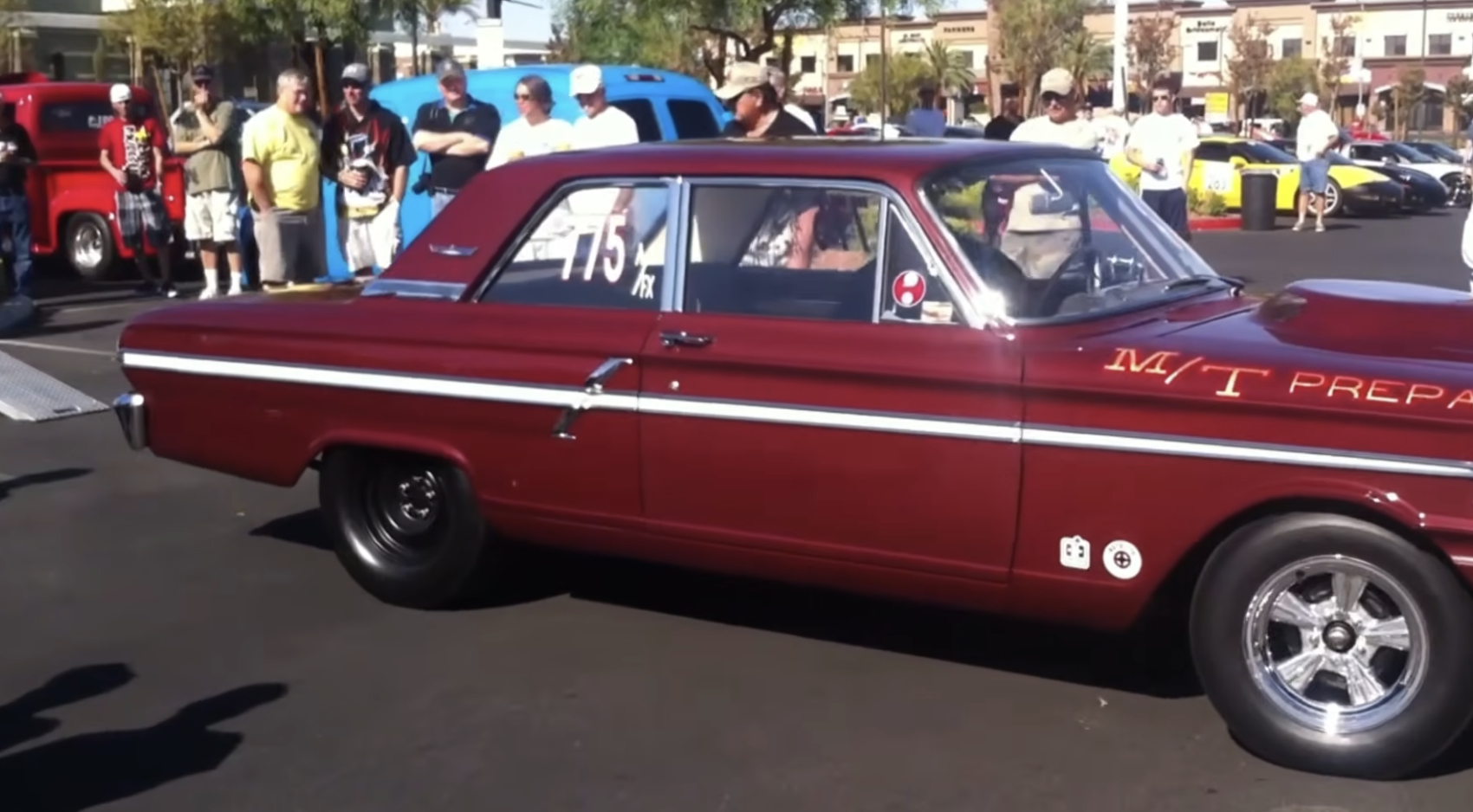 A maroon vintage car with racing decals is parked at a car show, surrounded by people standing and watching in a sunny outdoor lot.