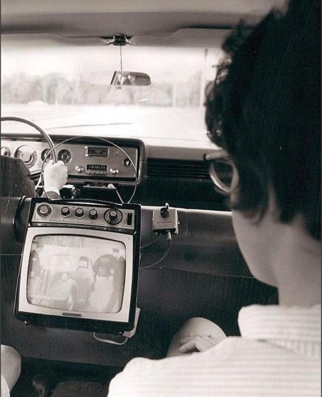 A black-and-white photo shows a person in the backseat of a car watching a small TV mounted on the front seat. The dashboard and steering wheel are visible, with a view out the windshield.