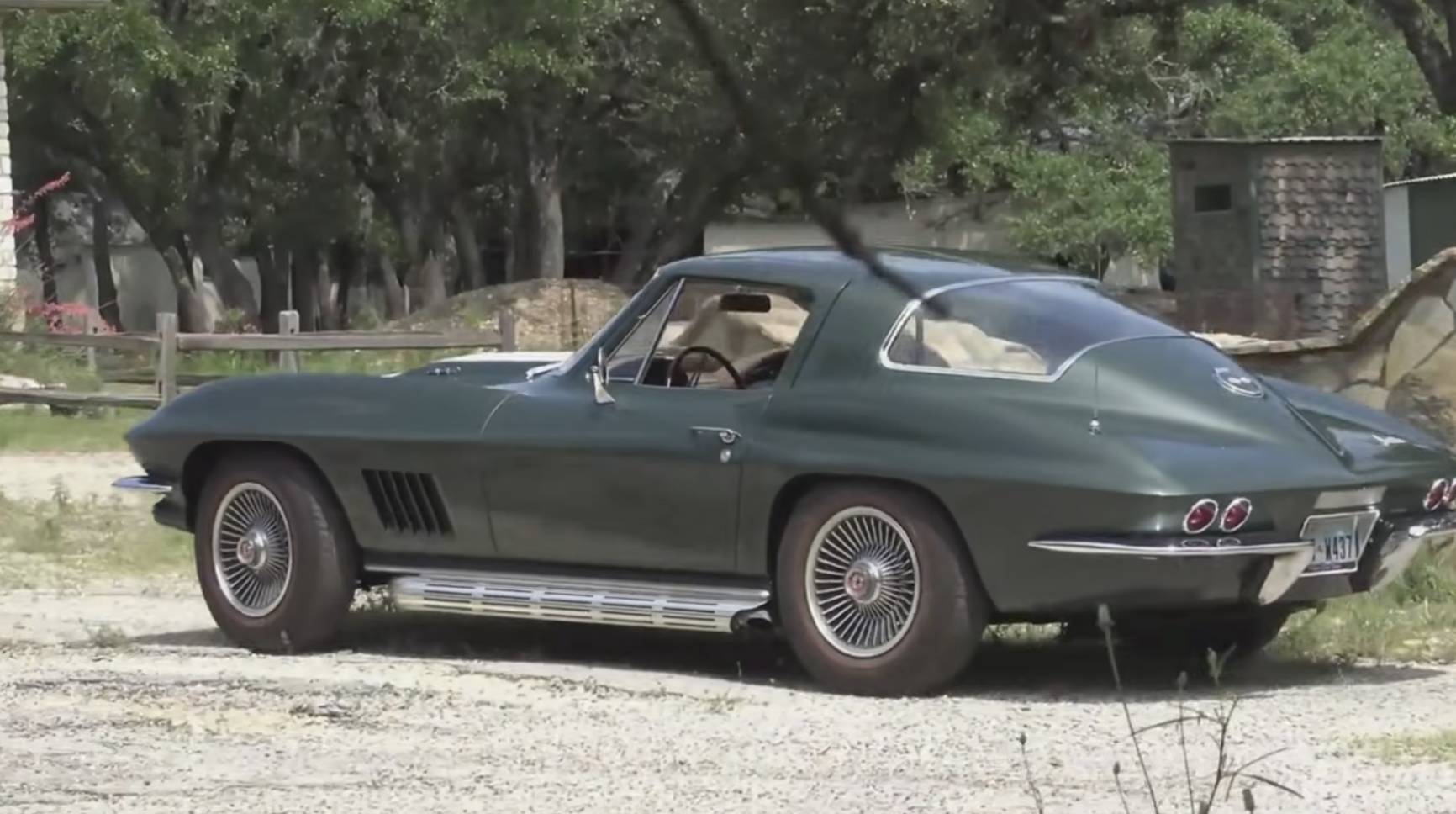 A classic dark green sports car with chrome accents and wire-spoke wheels is parked outdoors on a gravel surface, surrounded by trees and rustic buildings in the background.