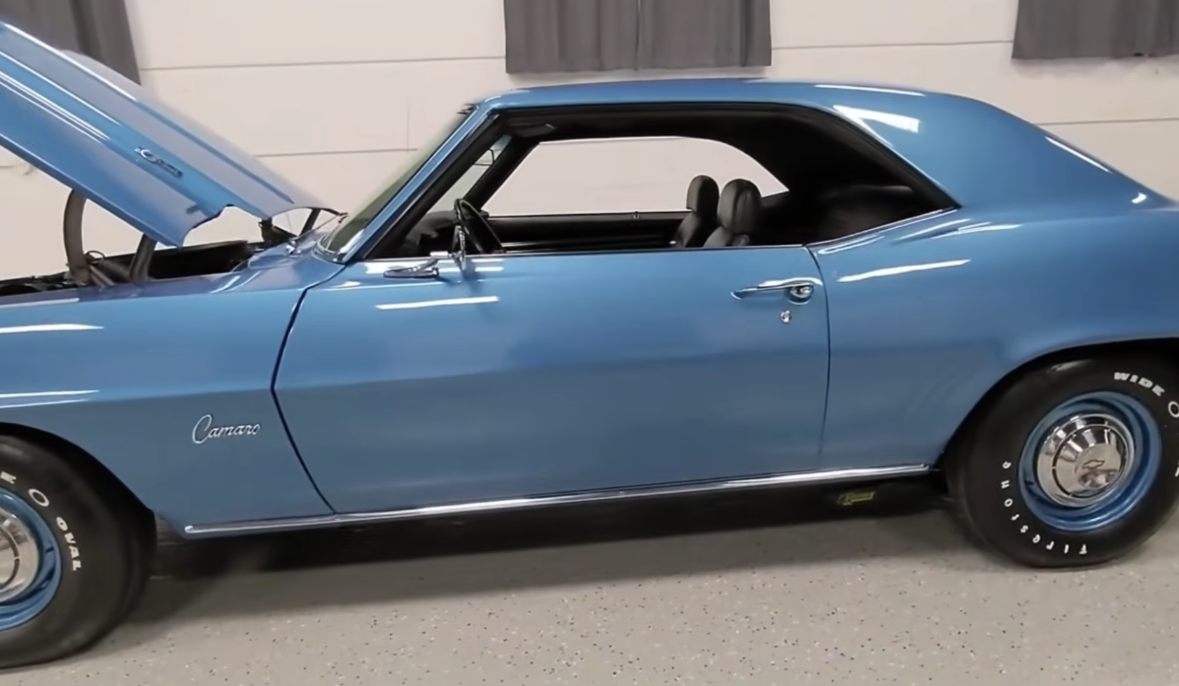 A classic blue Chevrolet Camaro with its hood open, parked indoors on a speckled floor, with gray curtains hanging on the white wall behind it.