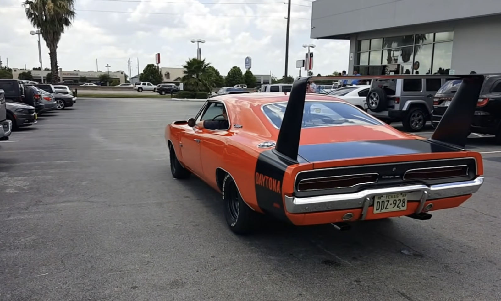 An orange classic Dodge Charger Daytona with a black rear wing and “Daytona” text on the side is parked outside a dealership, surrounded by other vehicles.