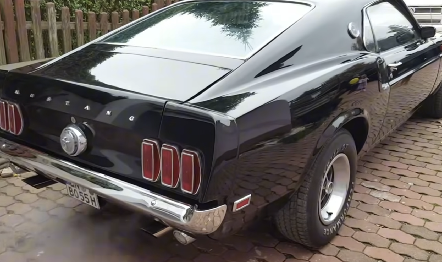 A black classic Ford Mustang is parked on a brick driveway, with its rear side visible. The car has chrome details and wide tires, and light exhaust smoke is coming from the tailpipe.