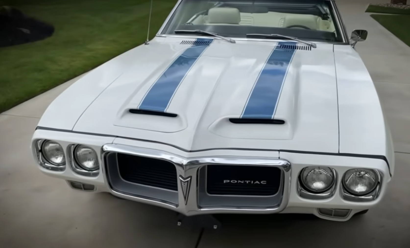 A classic white Pontiac muscle car with dual blue racing stripes parked on a concrete driveway next to green grass.