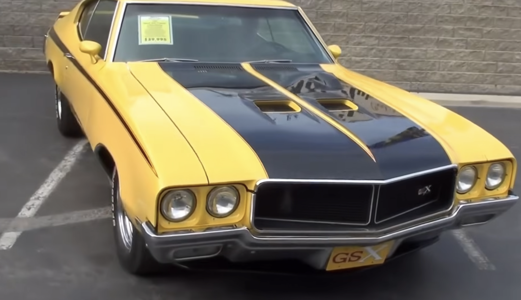 A yellow and black classic Buick GSX muscle car is parked on pavement near a stone wall, with a for-sale sign visible in the windshield.