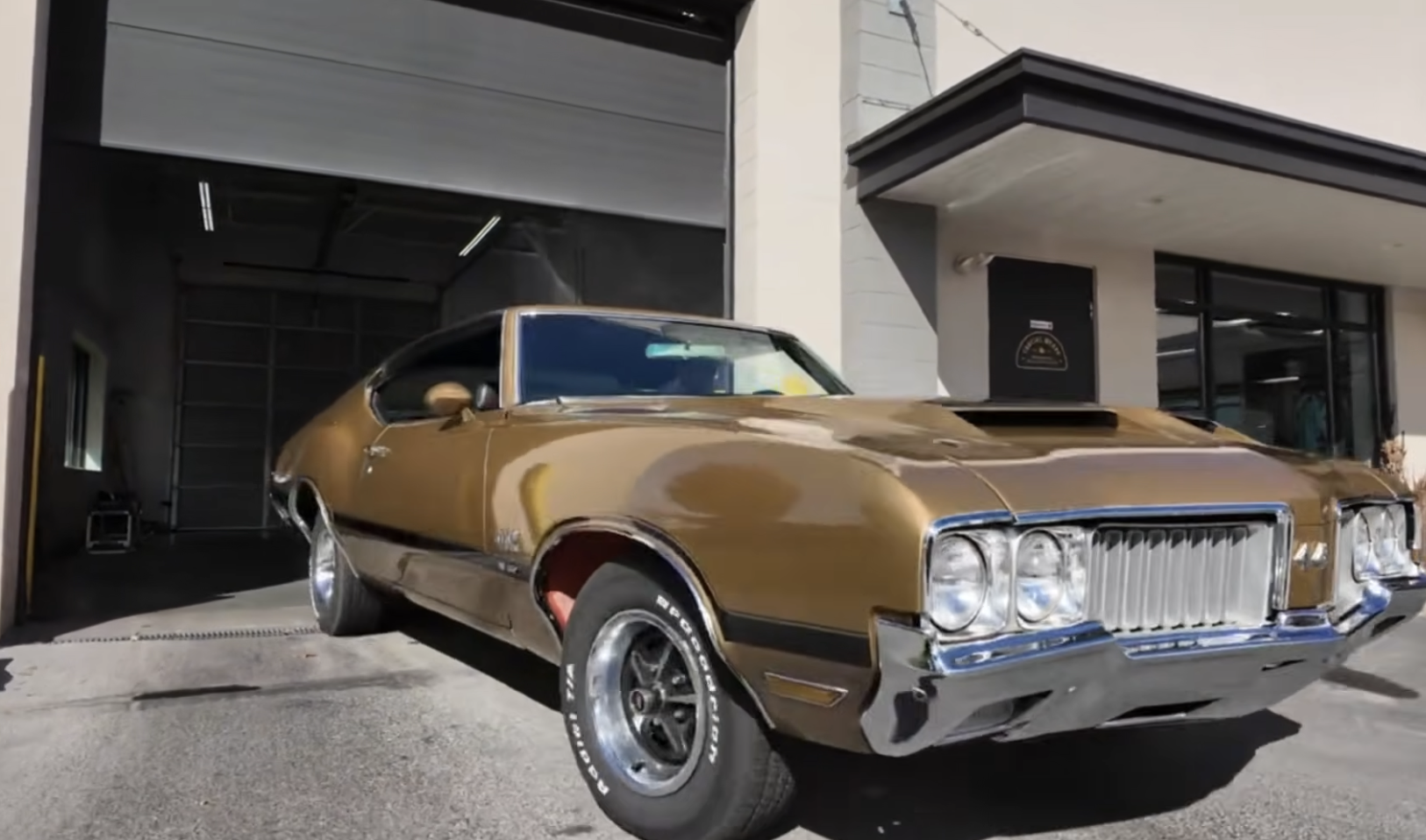 A vintage gold muscle car with chrome trim is exiting a garage with an open door on a sunny day. The building is beige with black accents.