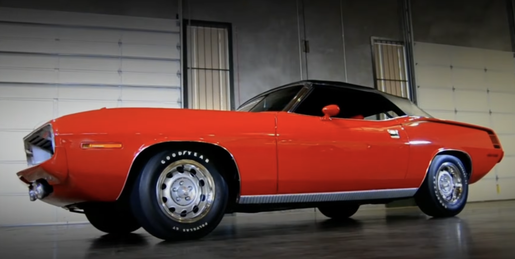 A red vintage muscle car with a black roof is parked indoors on a smooth floor, with a garage door and windows in the background.