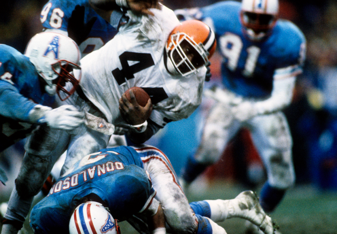 A football player in a white uniform runs with the ball while being tackled by multiple players in blue uniforms during a game. The players' uniforms are dirty, indicating rough play.