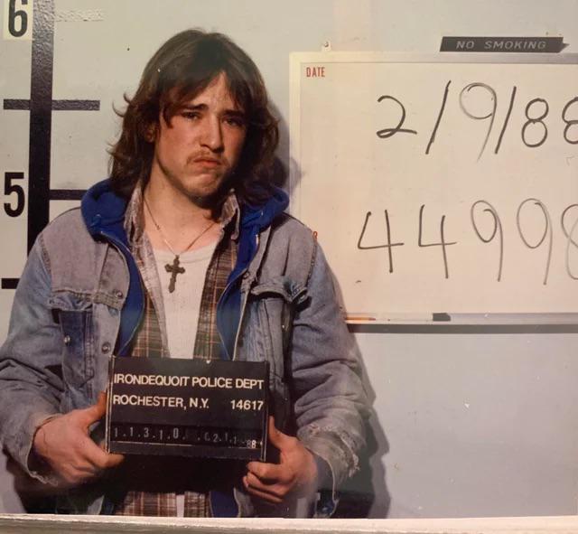 A young man with long hair, wearing a denim jacket and a cross necklace, poses for a mugshot holding a board labeled "Irondequoit Police Dept, Rochester, N.Y." A date and number are written on a whiteboard behind him.