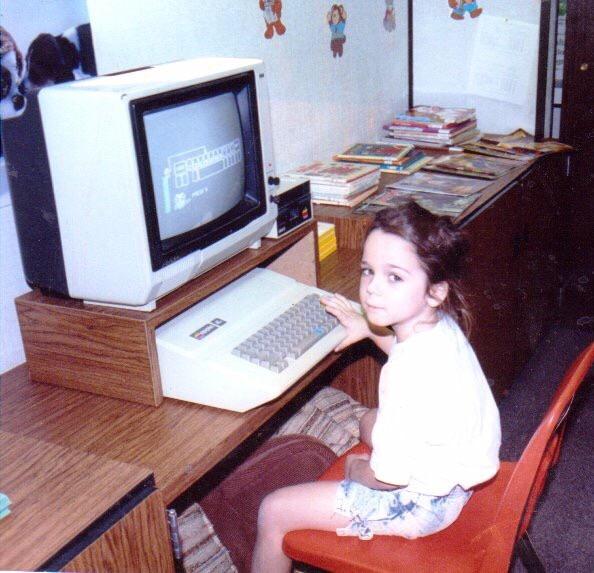 A young girl sits at a retro computer with a CRT monitor, surrounded by books and papers on a wooden desk. She looks back at the camera, her hand on the keyboard, while a game or program is displayed on the screen.