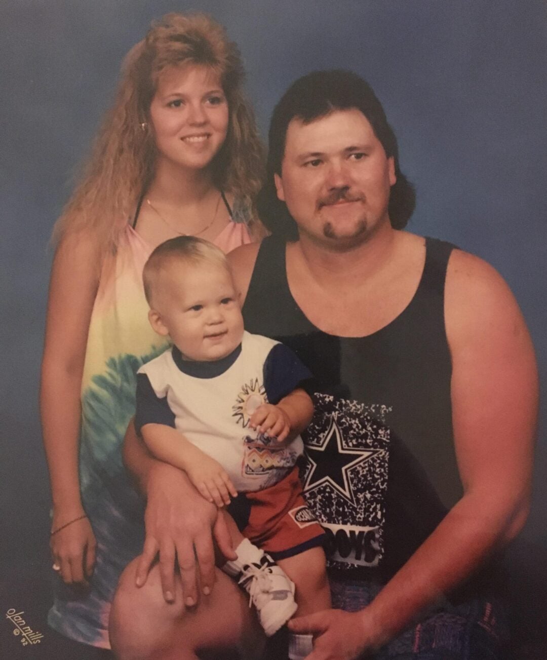 A woman with long, curly hair and a man with a mustache and mullet pose for a portrait, smiling, with a baby sitting on the man's lap. The family wears casual 1990s-style clothing against a blue background.