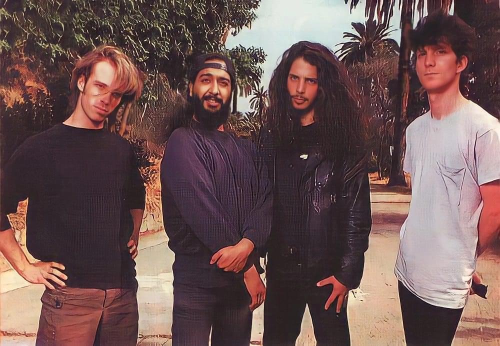 Four men stand outdoors on a sunny street lined with palm trees; they have casual poses and long hair, with one wearing a black leather jacket and the others in casual shirts and pants.