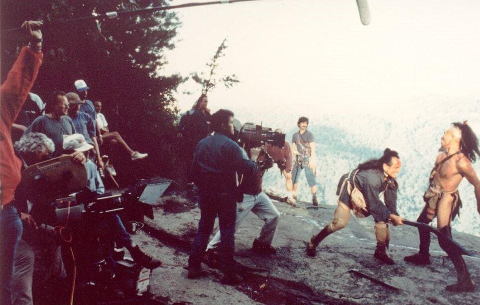 A film crew records a dramatic fight scene between two actors in costume on a rocky cliff, with cameras, crew members, and a boom microphone visible in the shot. Forested hills are in the background.