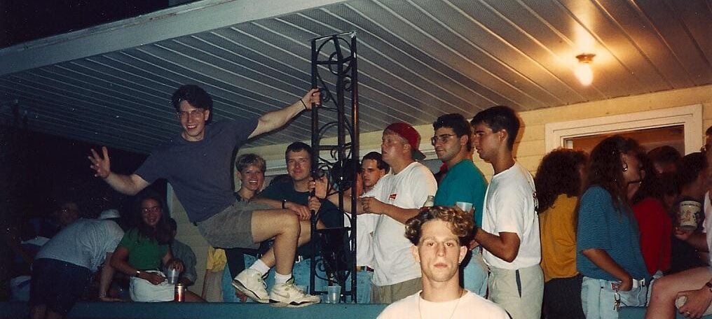 A group of young people gather on a porch at night, many smiling and talking. One person poses playfully on the railing, while another stands in front wearing a "Sweaty Nipples" band shirt. The scene looks lively and social.