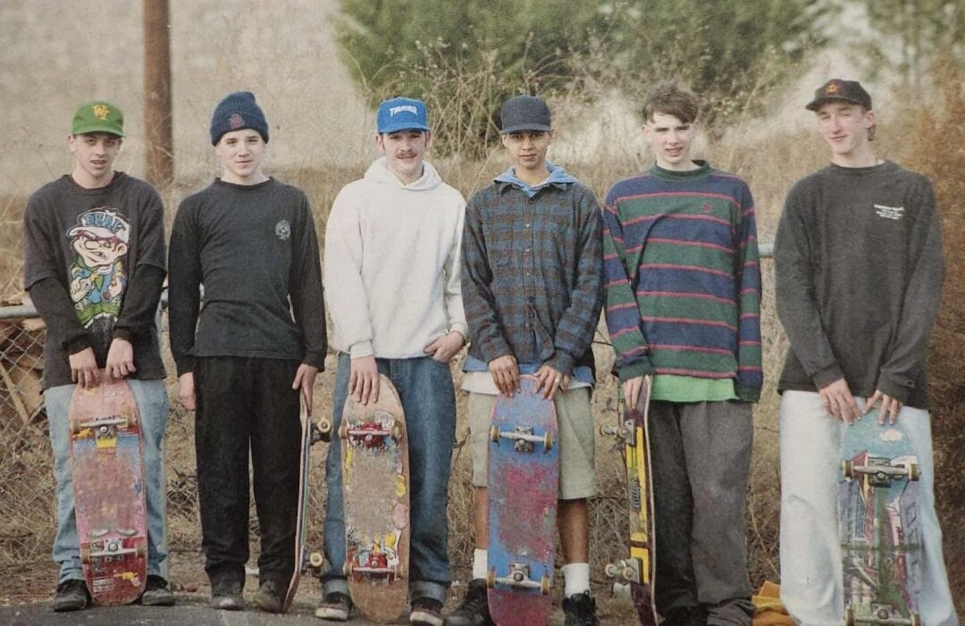 Six teenage boys stand outdoors in a row, each holding a skateboard vertically. They wear casual 1990s-style clothing including beanies, caps, and sweatshirts, and stand on a paved surface with dry grass and trees behind them.