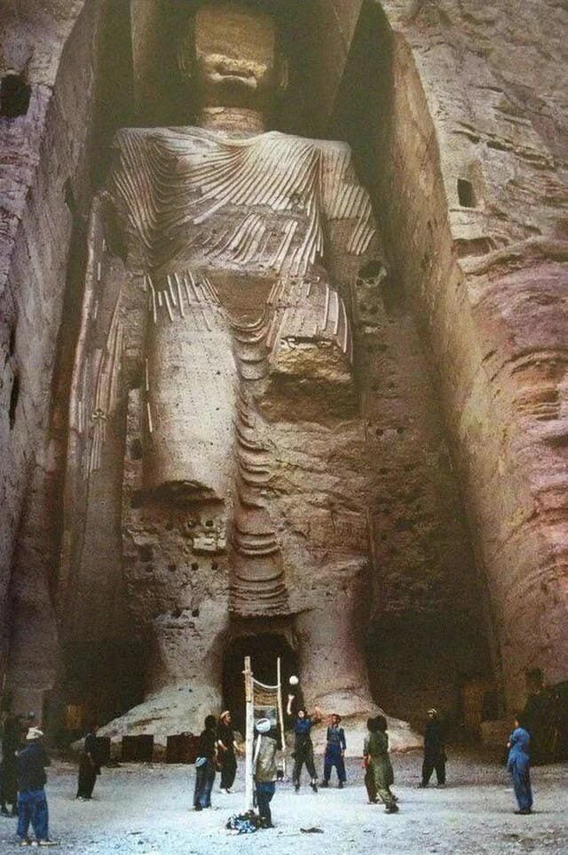 A group of people stand at the base of a giant, ancient stone Buddha statue carved into a cliffside, showing the statue’s massive scale and intricate details, with scaffolding and equipment on the ground.