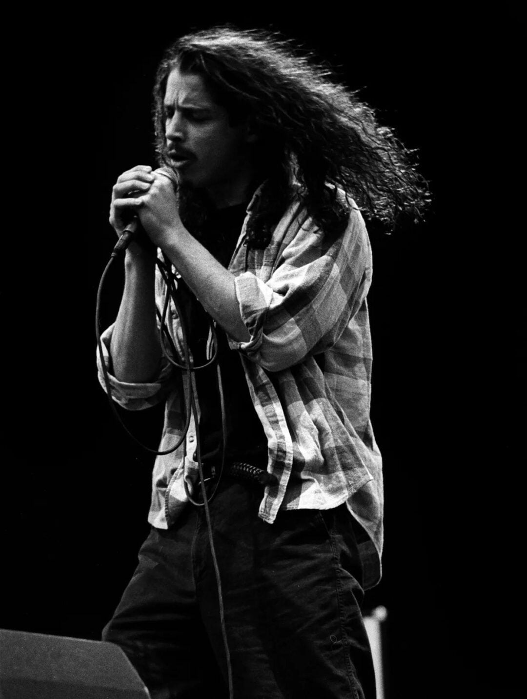A man with long curly hair sings passionately into a microphone on stage, wearing a plaid shirt over a dark t-shirt and cargo pants, against a dark background.