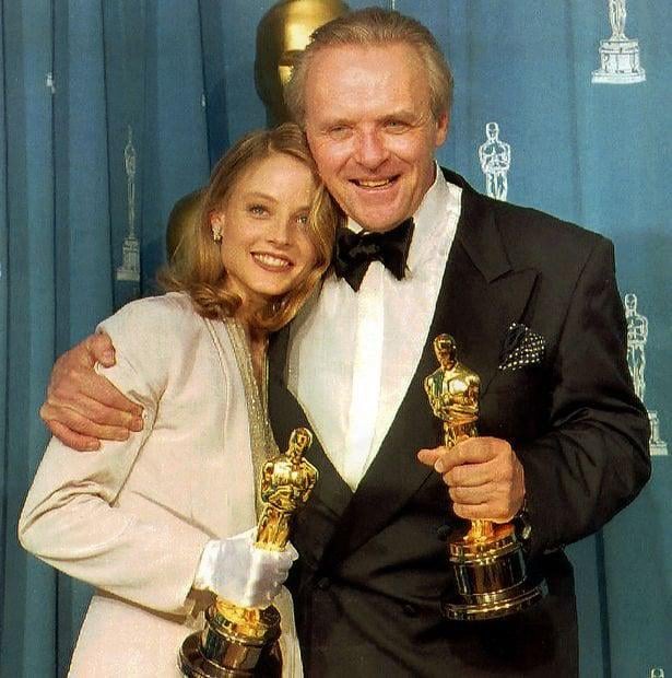 A woman and a man in formal attire smile and pose together, each holding an Oscar trophy. Blue curtain with Oscar statuette logos is in the background. The man has his arm around the woman.