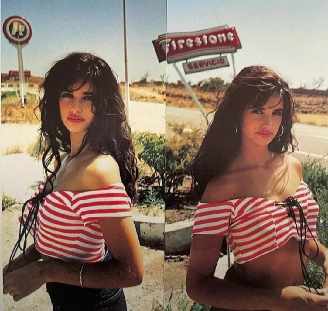 A woman with long dark hair, wearing a red and white striped off-shoulder top, stands outdoors near a road. Behind her are dry grass, a Firestone service sign, and a no-entry traffic sign.